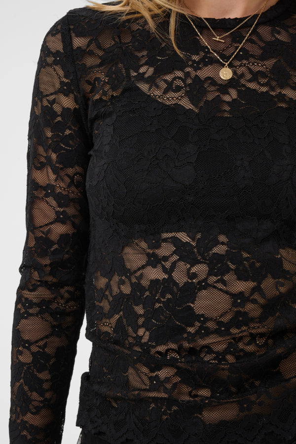 A close-up of a person wearing a black lace long-sleeve top, showing the intricate floral lace pattern and sheer mesh details. The person is also wearing layered gold necklaces.