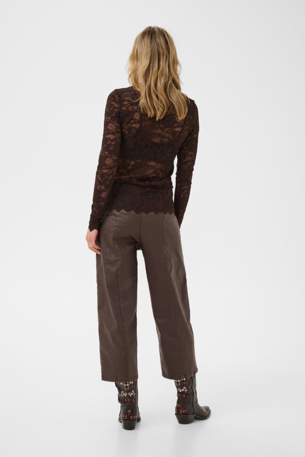 A woman viewed from behind wearing a dark brown lace long-sleeve top, brown wide-leg cropped pants, and dark patterned ankle boots against a light gray studio background.