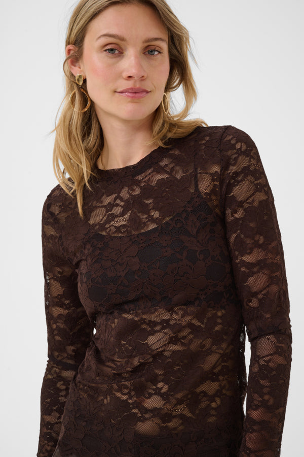 A woman wearing a dark brown lace long-sleeve top against a plain light gray studio background.