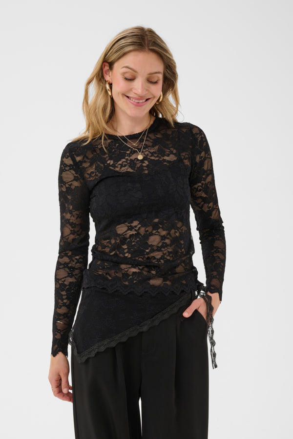A woman wearing a black lace long-sleeve top with a scalloped hem and black pants, photographed against a plain light gray studio background.