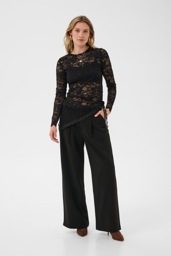 A woman modeling a black long-sleeve top with a sheer lace overlay and black wide-leg trousers, standing against a plain light gray studio background.
