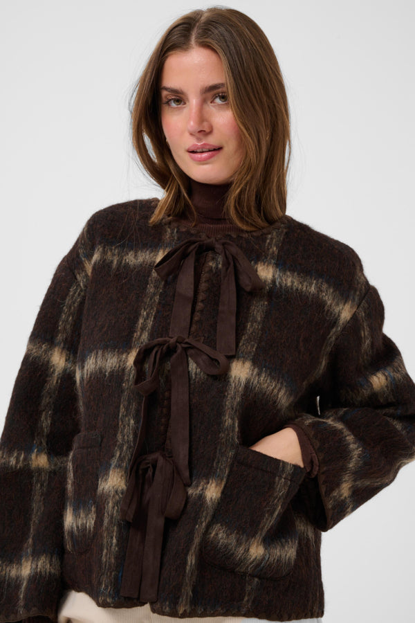 A woman wearing a dark brown and beige plaid cropped jacket with a high neck and front tie detail, photographed against a light gray studio background.