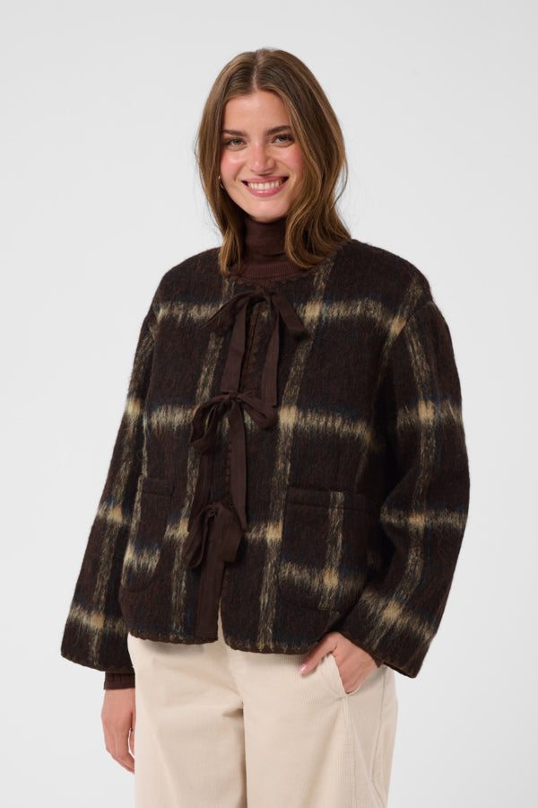 A woman wearing a dark brown and beige plaid cropped jacket with a tie-neck detail, paired with cream trousers, against a plain light gray background.
