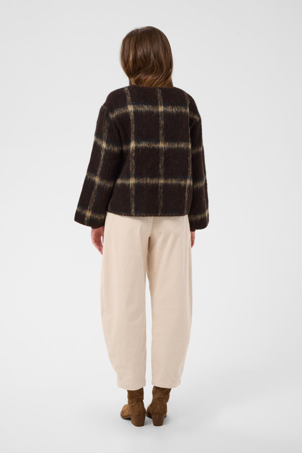 A model shown from the back wearing a dark brown and beige plaid cropped jacket, cream-colored baggy pants, and brown suede boots, set against a plain light gray studio background.