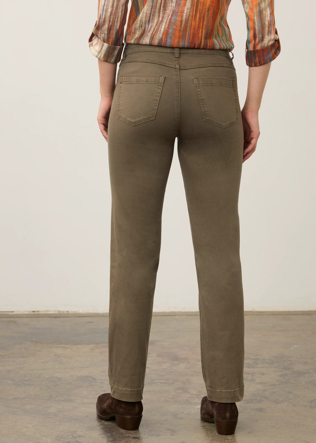 A person shown from the back wearing straight-leg olive green jeans, a multicolor striped shirt, and brown shoes, standing against a plain off-white wall and concrete floor.