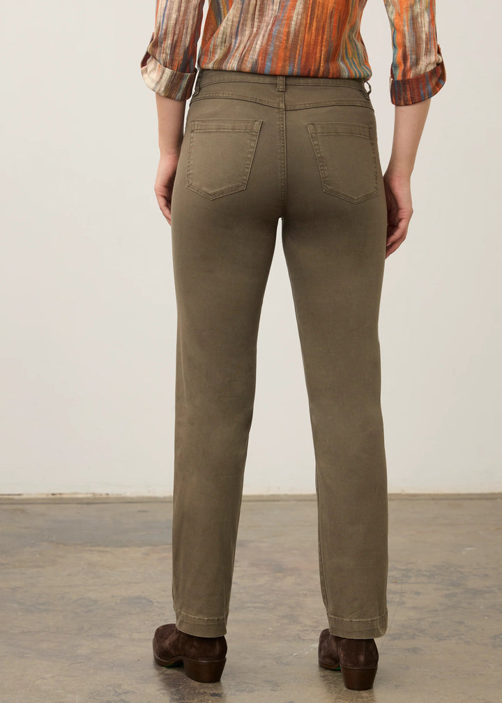 A person shown from the back wearing straight-leg olive green jeans, a multicolor striped shirt, and brown shoes, standing against a plain off-white wall and concrete floor.