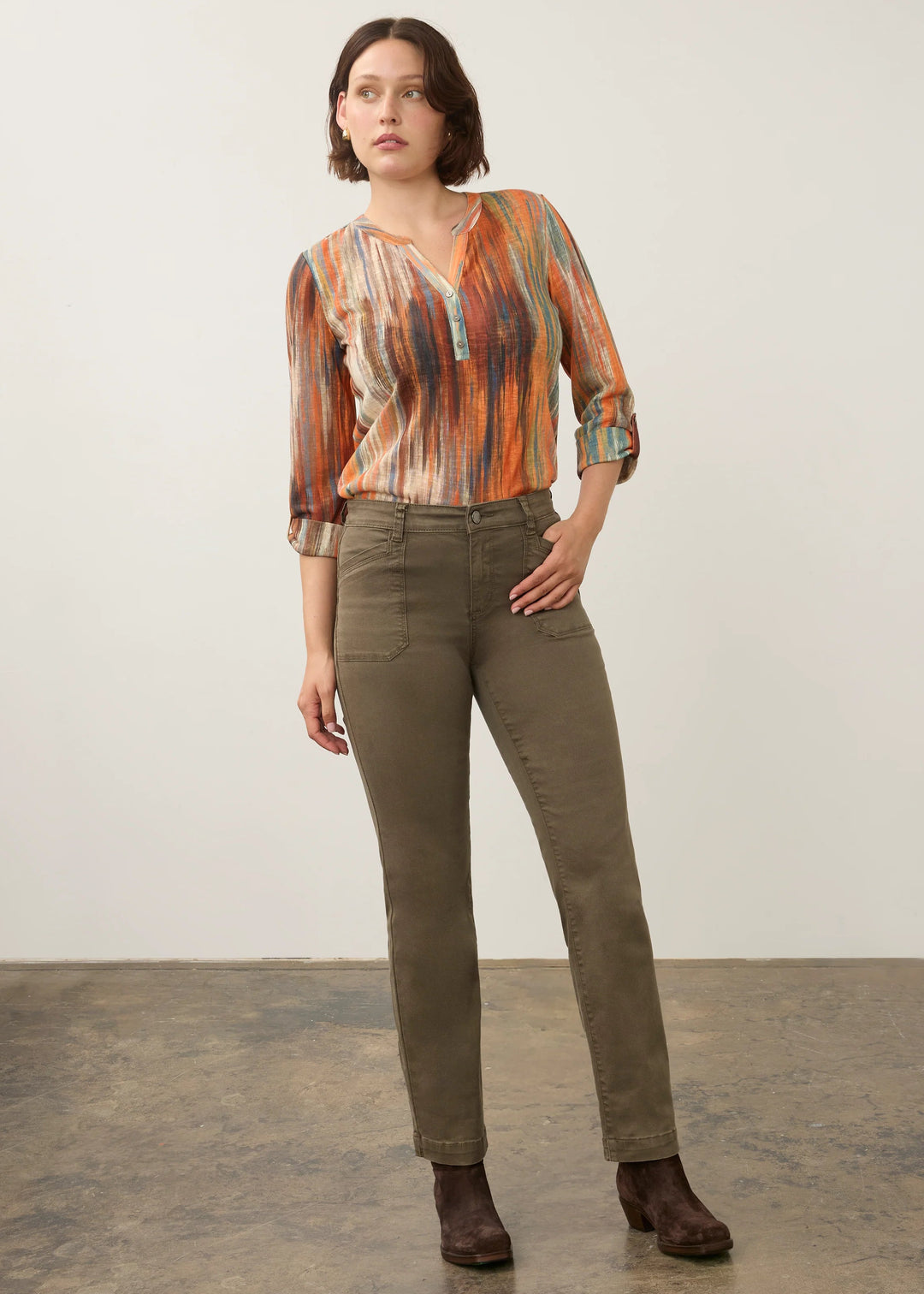 A woman modeling a long-sleeve button-up shirt with an abstract orange, brown, and beige pattern, paired with olive green trousers and dark brown ankle boots, standing against a neutral studio backdrop.