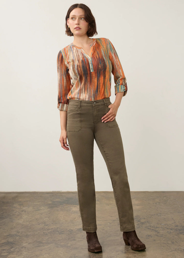 A woman modeling a long-sleeve button-up shirt with an abstract orange, brown, and beige pattern, paired with olive green trousers and dark brown ankle boots, standing against a neutral studio backdrop.