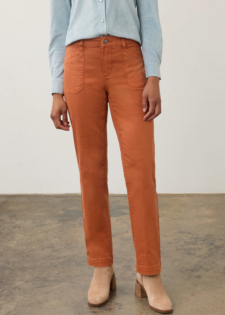 A model wearing a pair of rust-orange slim-fit jeans, paired with a light blue button-down shirt and beige ankle boots, standing on a concrete floor against a white wall.