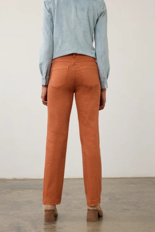 A person viewed from behind wearing straight-leg orange-brown trousers, a light blue long-sleeve top, and brown shoes in a minimalist studio setting with a concrete floor and off-white wall.