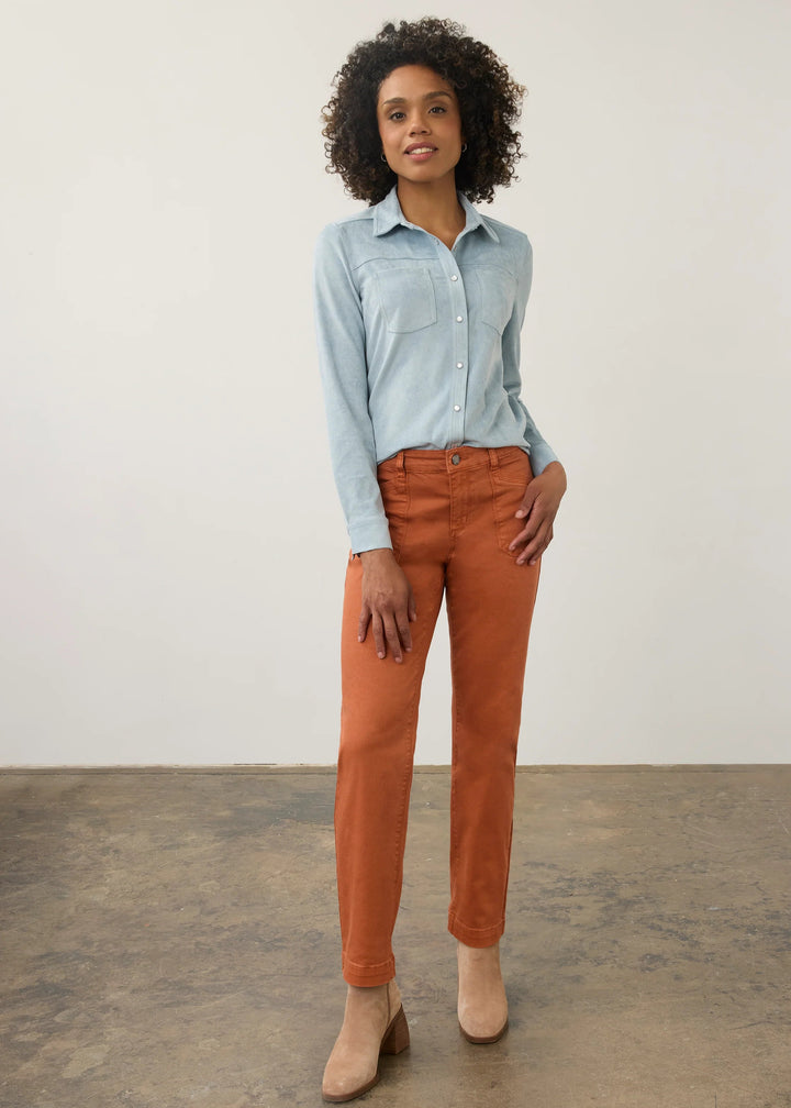 A woman stands in a studio wearing a light blue long-sleeve button-up shirt, rust-orange cropped pants, and beige ankle boots against a plain off-white wall and concrete floor.