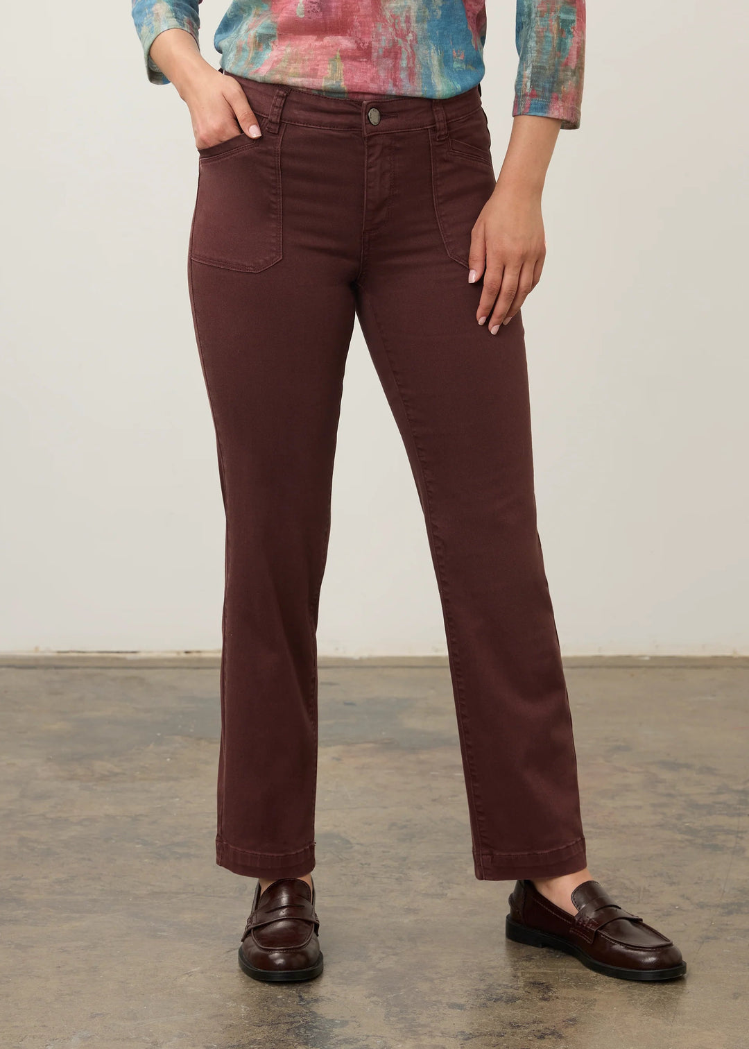 A model wearing a pair of dark brown straight-leg jeans, paired with a colorful abstract print long-sleeve shirt and dark brown leather loafers, standing against a plain light gray wall.