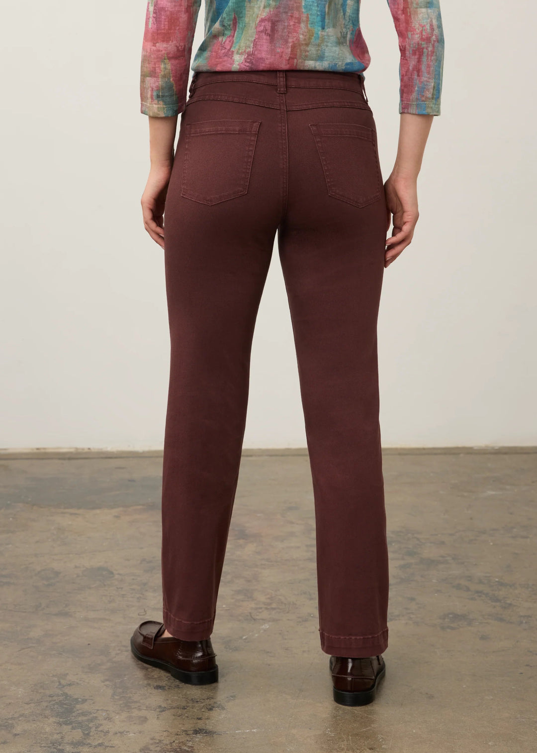 A person shown from the back wearing slim-fit burgundy trousers, a multicolored abstract print long-sleeve top, and dark brown leather loafers, standing against a plain off-white wall and concrete floor.