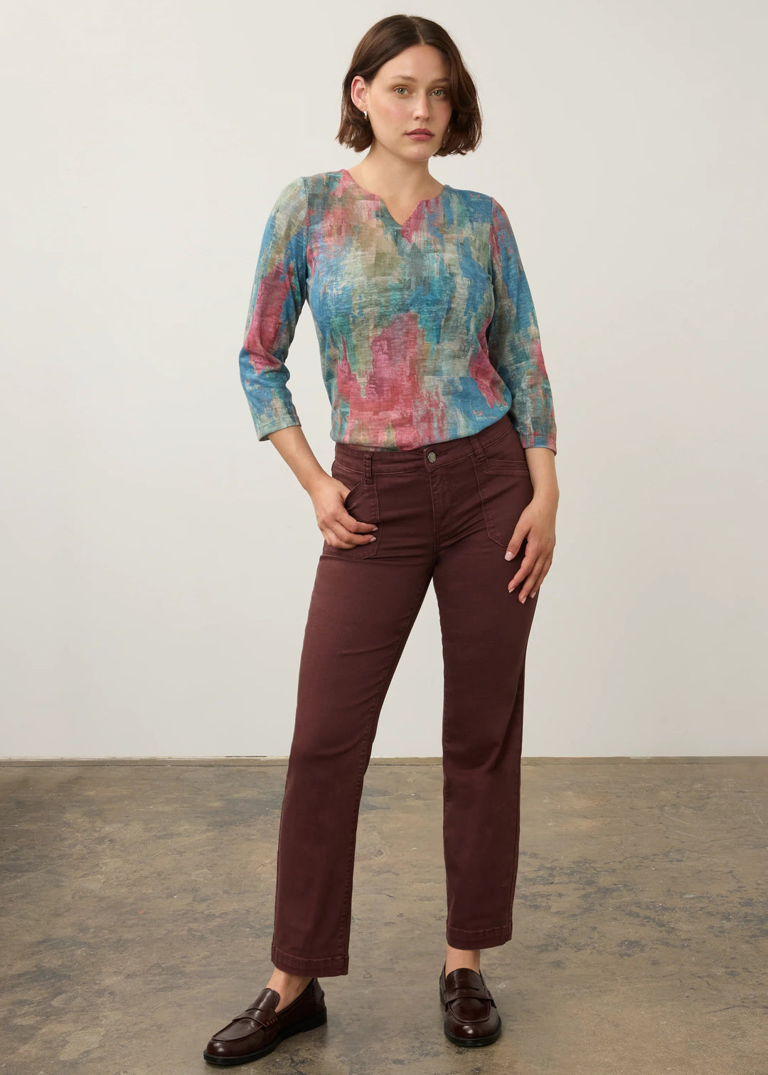 A woman modeling a three-quarter sleeve V-neck blouse with an abstract blue, pink, and teal pattern, paired with dark brown trousers and dark brown leather loafers, standing against a neutral studio backdrop.