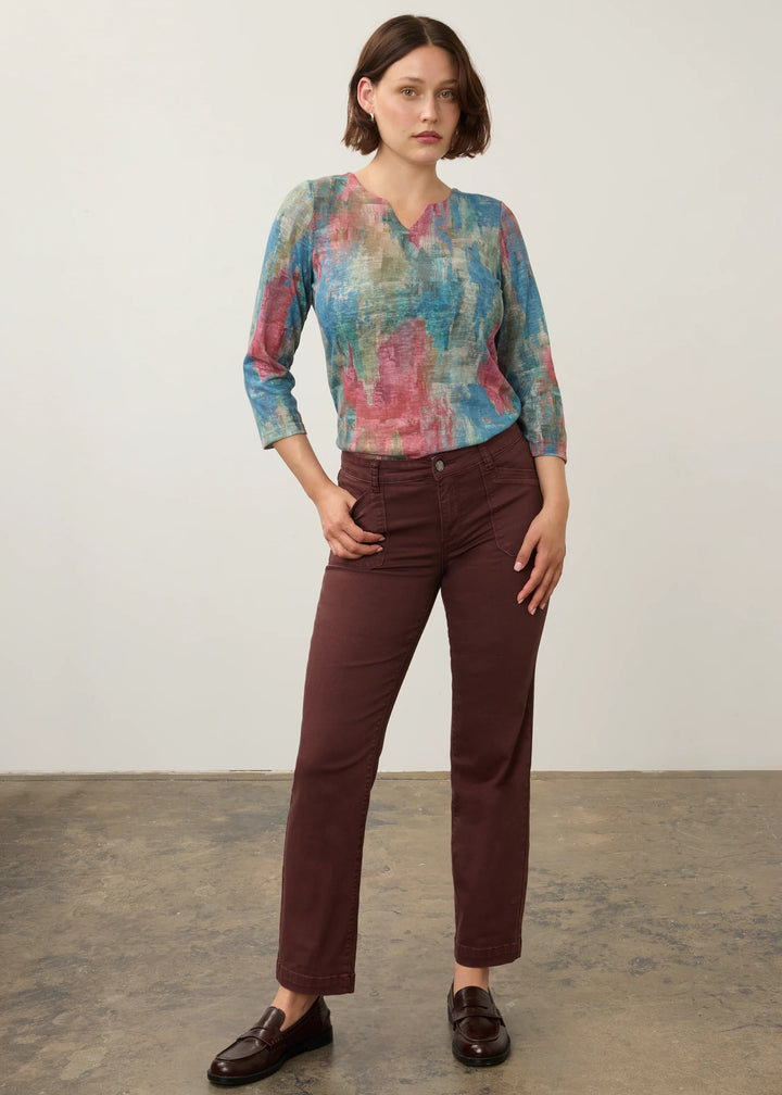 A woman modeling a three-quarter sleeve V-neck blouse with an abstract blue, pink, and teal pattern, paired with dark brown trousers and dark brown leather loafers, standing against a neutral studio backdrop.