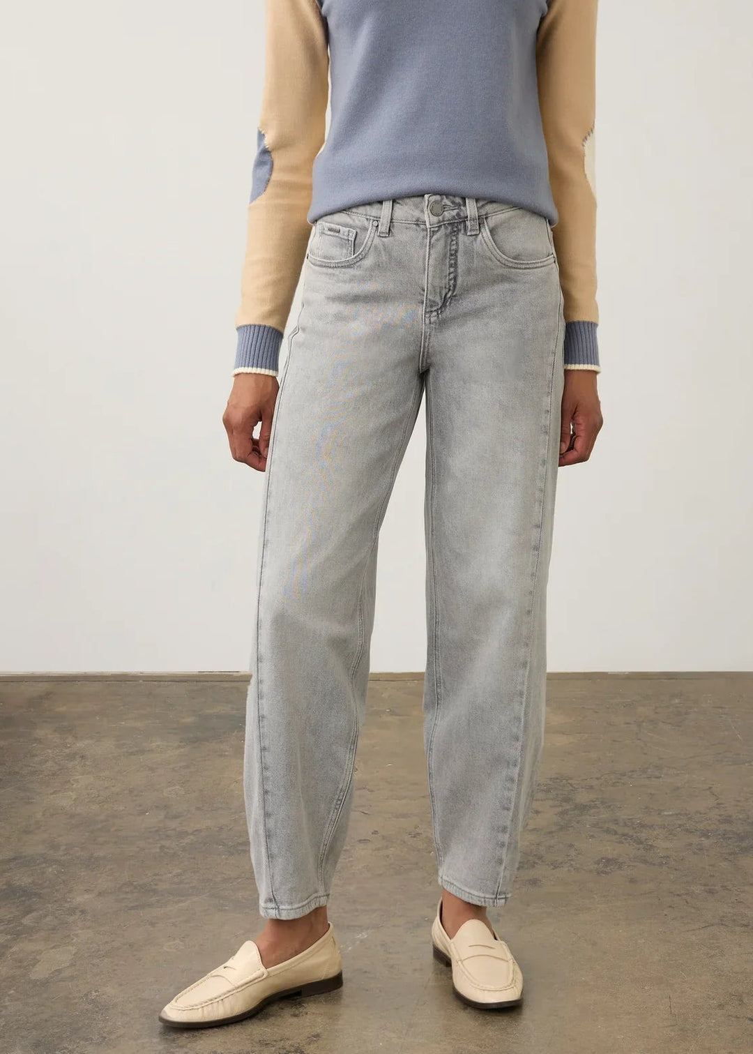 A model wearing a light blue-gray colorblock raglan sweater with beige sleeves, light-wash straight-leg jeans, and cream loafers, standing against a neutral studio background.