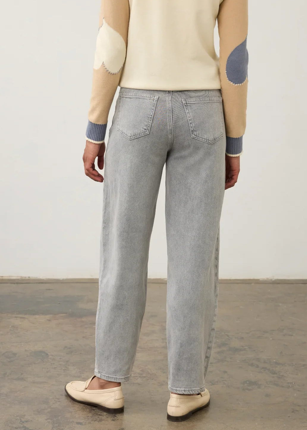 A person shown from the back wearing light gray wide-leg jeans, a cream sweater with beige and blue patchwork elbow patches, and beige slip-on shoes, standing against a plain light wall and concrete floor.