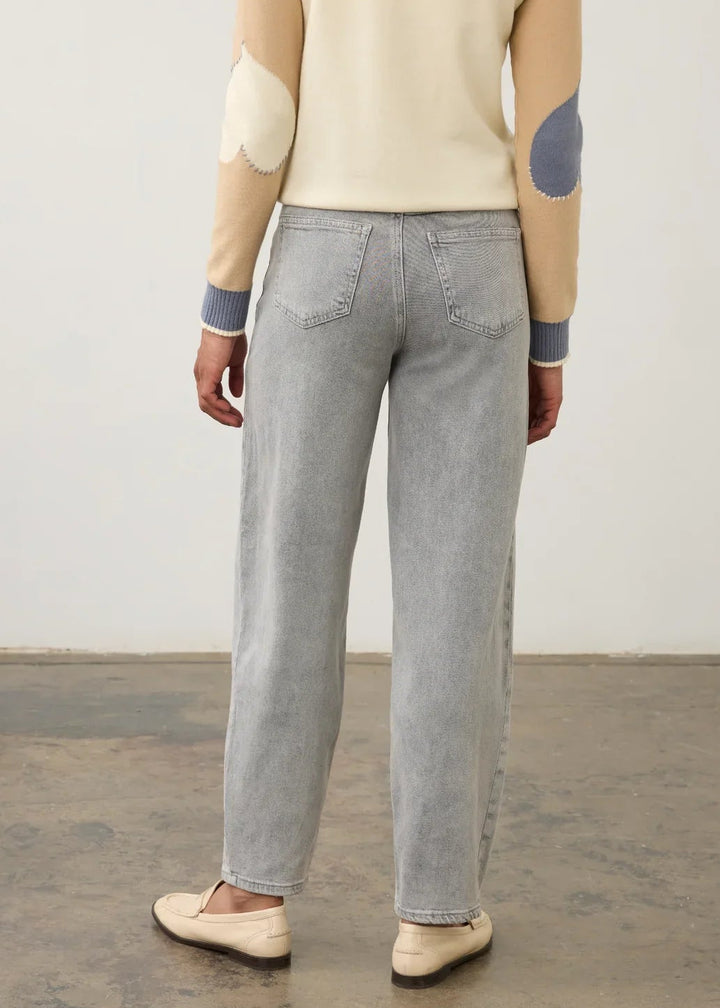 A person shown from the back wearing light gray wide-leg jeans, a cream sweater with beige and blue patchwork elbow patches, and beige slip-on shoes, standing against a plain light wall and concrete floor.