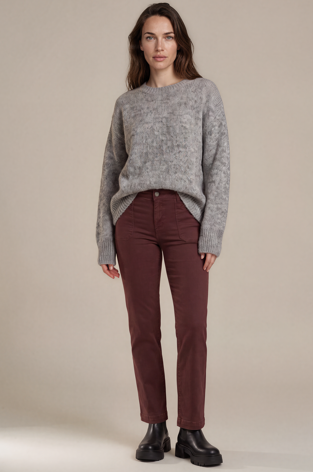 A woman stands against a plain beige studio background wearing a heather gray knit sweater, dark burgundy pants, and black boots.
