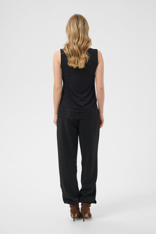 A woman viewed from behind wearing a black sleeveless top and black wide-leg pants, standing against a light gray studio background.