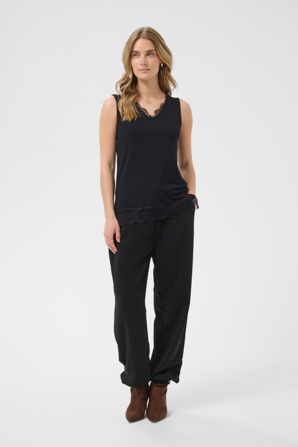 A woman modeling a black sleeveless top with a ruffled hem and matching black wide-leg pants, paired with brown ankle boots, against a light gray studio background.