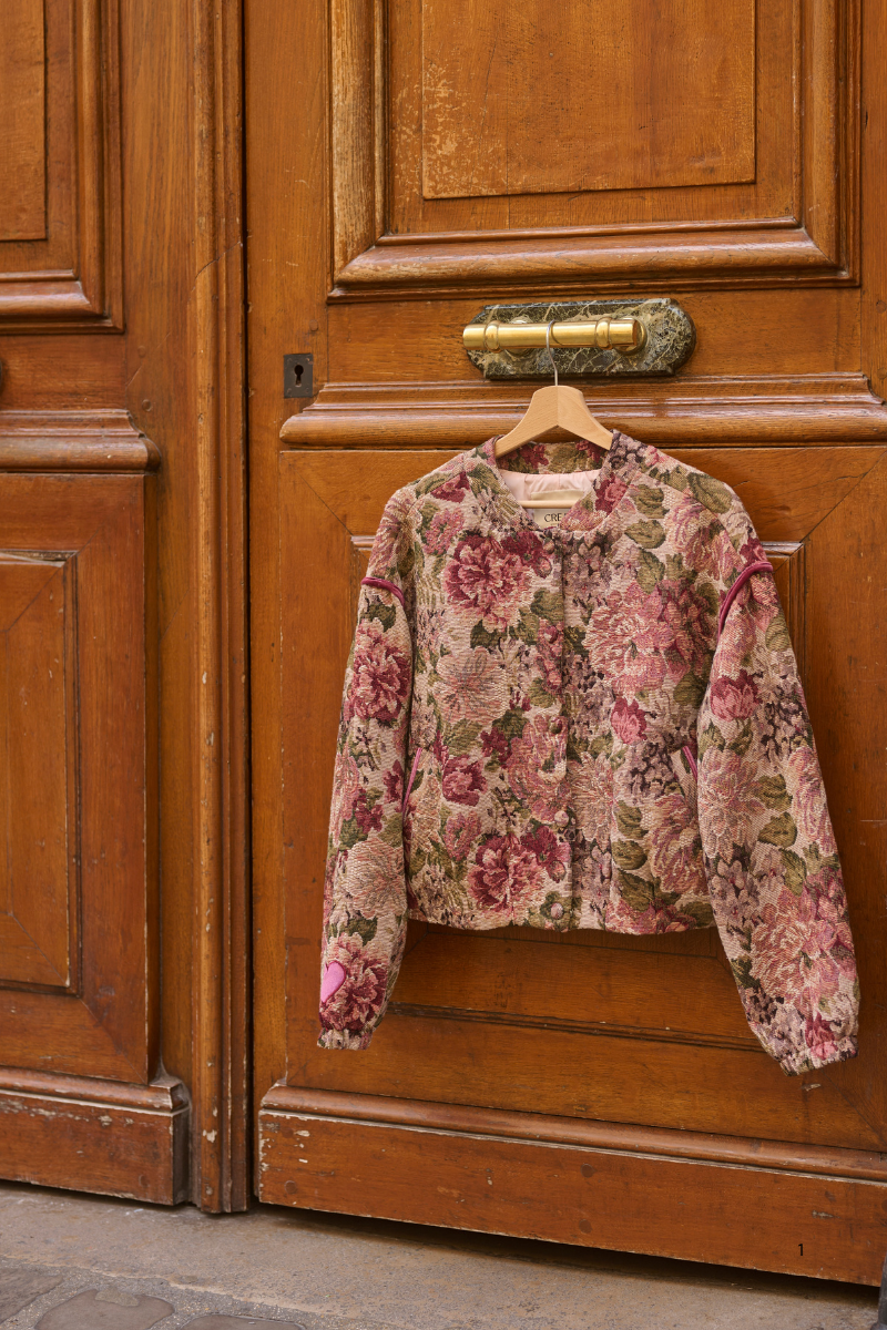 A cropped long-sleeve floral jacket with a pink, red, and green rose pattern hangs on a wooden hanger against a rustic brown wooden door.