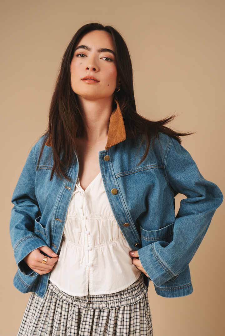 A woman modeling a cropped blue denim jacket with a tan corduroy collar, worn over a white blouse and a black-and-white plaid skirt, against a solid beige studio background.