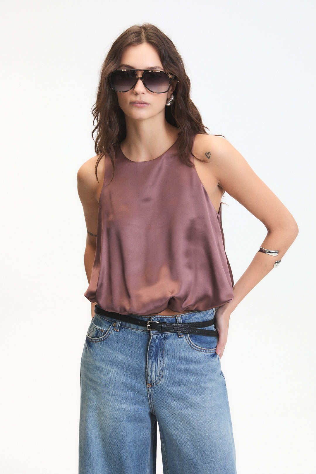 Woman wearing a brown sleeveless top and blue jeans with sunglasses on a white background