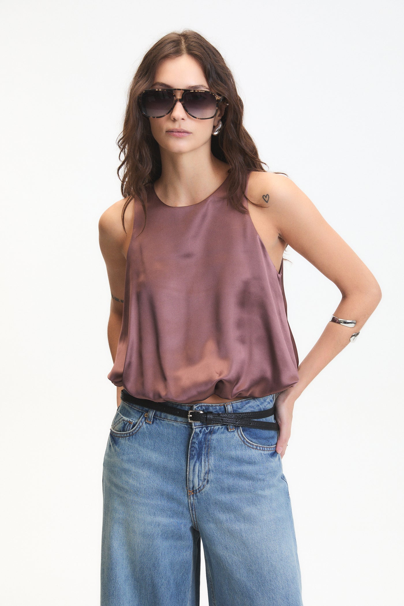Woman wearing a brown sleeveless top and blue jeans with sunglasses on a white background