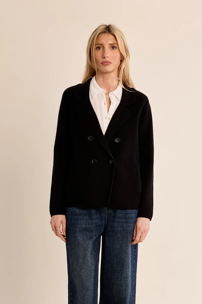 A woman modeling a black double-breasted blazer over a white collared shirt and blue jeans, standing against a plain beige studio background.