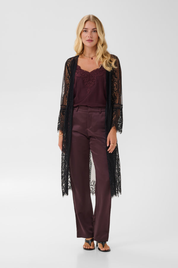 A blonde model stands against a light gray studio background wearing a dark plum satin pantsuit with a matching lace camisole and a sheer black lace open-front cardigan.