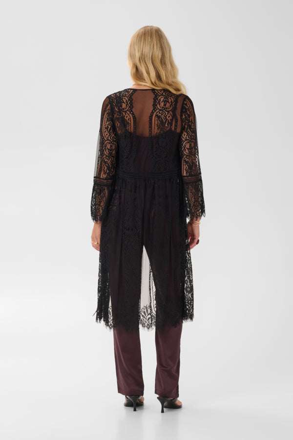 A woman viewed from the back wearing a black sheer lace long tunic over a dark dress, paired with mauve-burgundy pants and black heels, against a light gray studio background.