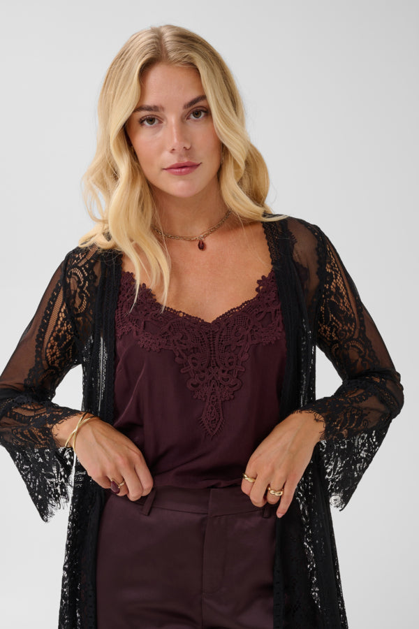 A blonde woman models a black sheer lace kimono cardigan over a deep plum purple lace camisole and matching trousers, set against a plain light gray studio background.