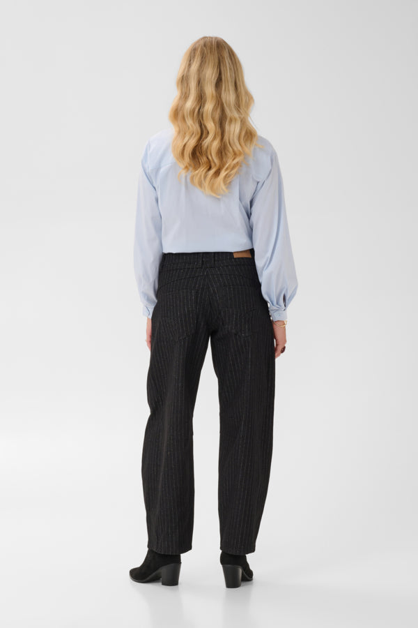 A woman viewed from behind wearing a light blue long-sleeve blouse and dark pinstriped trousers, standing against a plain light gray studio background.
