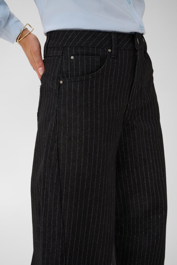 Close-up of a person wearing black wide-leg trousers with thin white pinstripes, paired with a light blue shirt. The photo focuses on the waist, front pockets, and fabric texture against a plain light gray background.