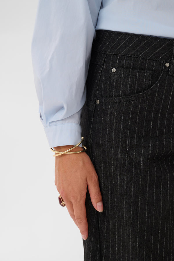 Close-up of a person wearing dark charcoal pinstriped trousers and a light blue long-sleeve shirt, with a gold wrap bracelet on the wrist against a plain light background.