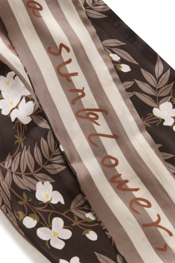 A close-up of a patterned scarf featuring vertical stripes in taupe and cream, overlaid with a dark brown floral print of white flowers and leaves. The word 'sunflower' is printed in brown script across the center.