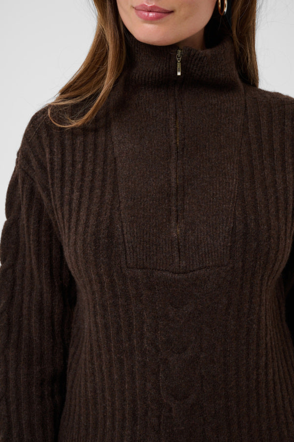 A close-up of a woman wearing a dark brown ribbed knit mock-neck sweater with a half-zip closure at the collar.