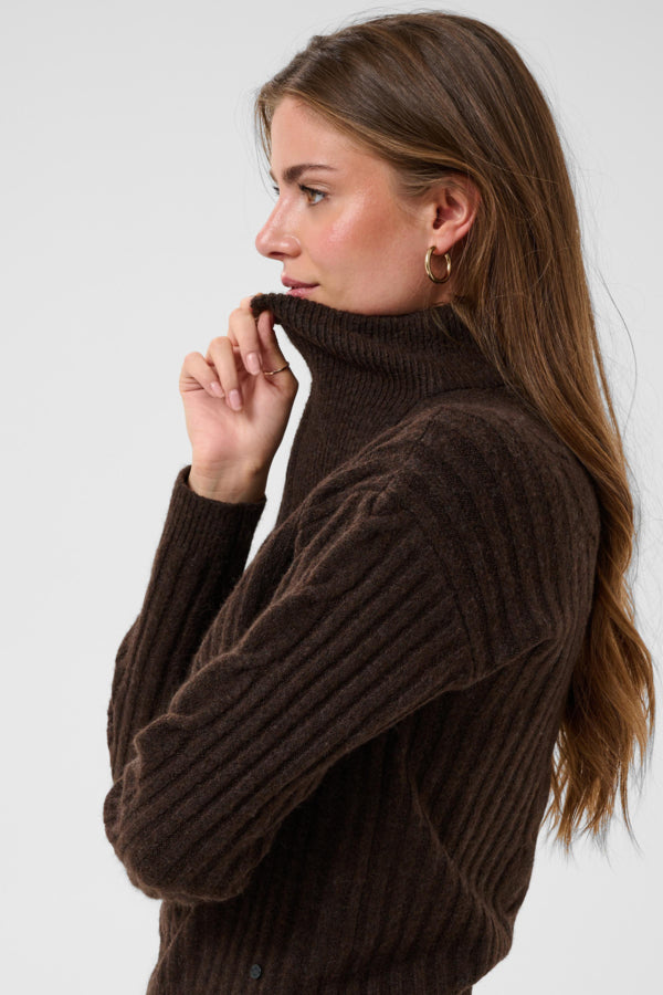 A woman wearing a dark brown ribbed turtleneck sweater, shown in profile against a light gray studio background.