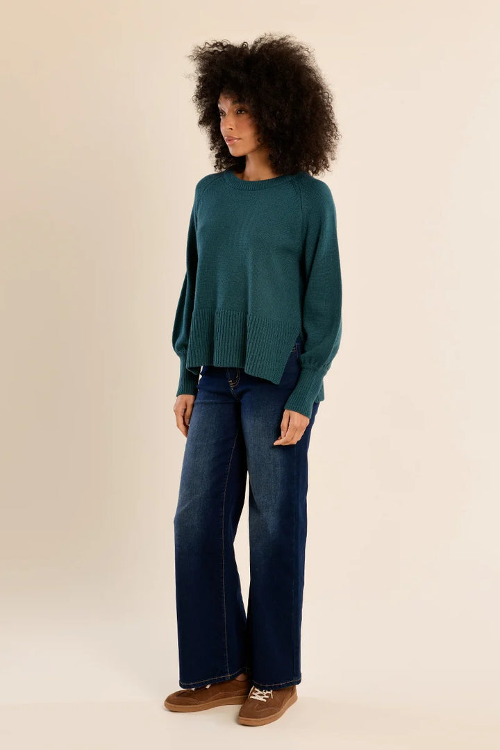 A model with curly dark hair stands in a studio wearing a teal-green long-sleeve knit sweater, dark blue wide-leg jeans, and brown suede shoes against a warm beige background.