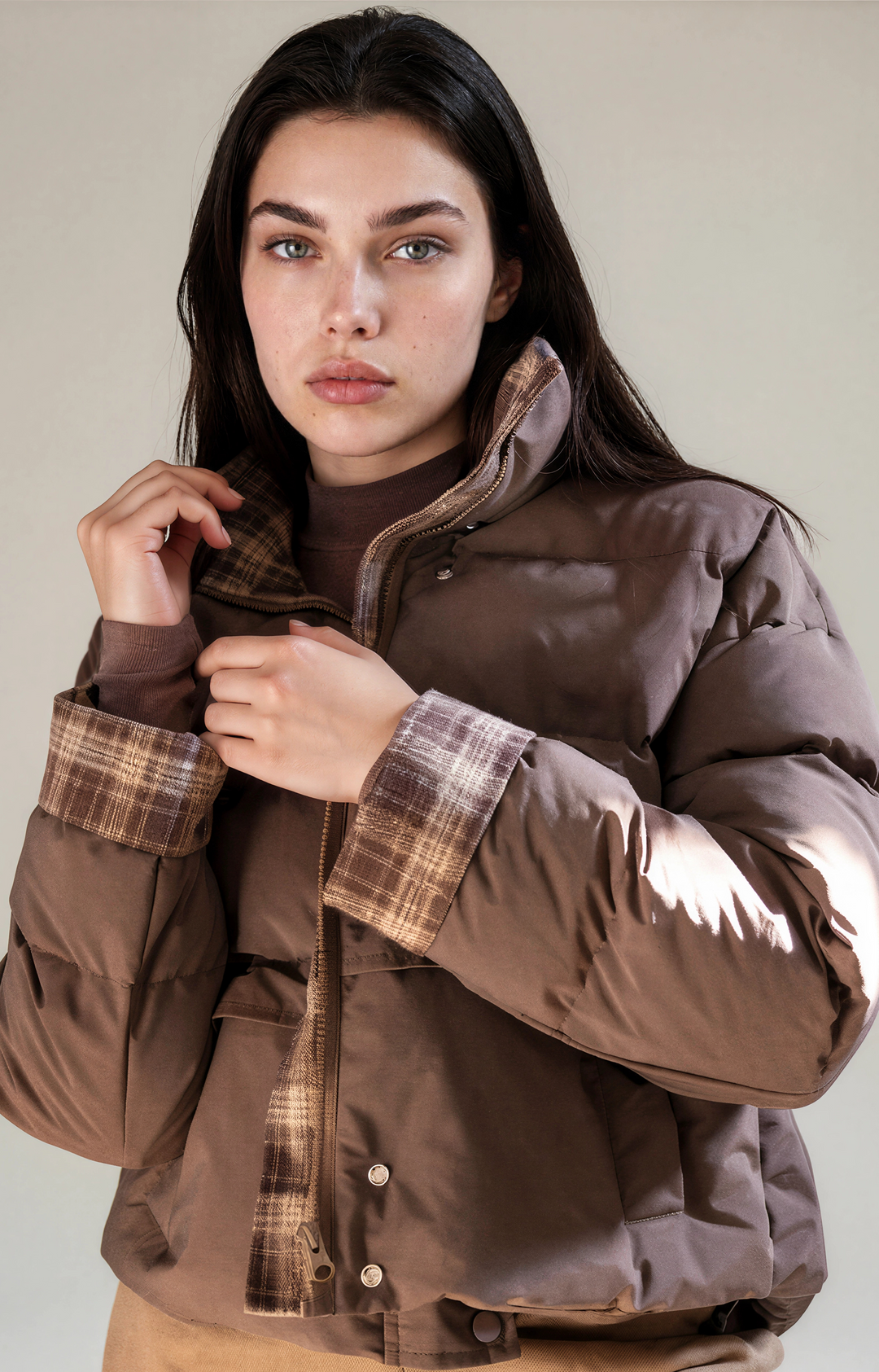 A model wearing a brown puffer jacket with a brown plaid lining on the collar, cuffs, and front placket, paired with a brown turtleneck and tan trousers, set against a plain beige studio background.