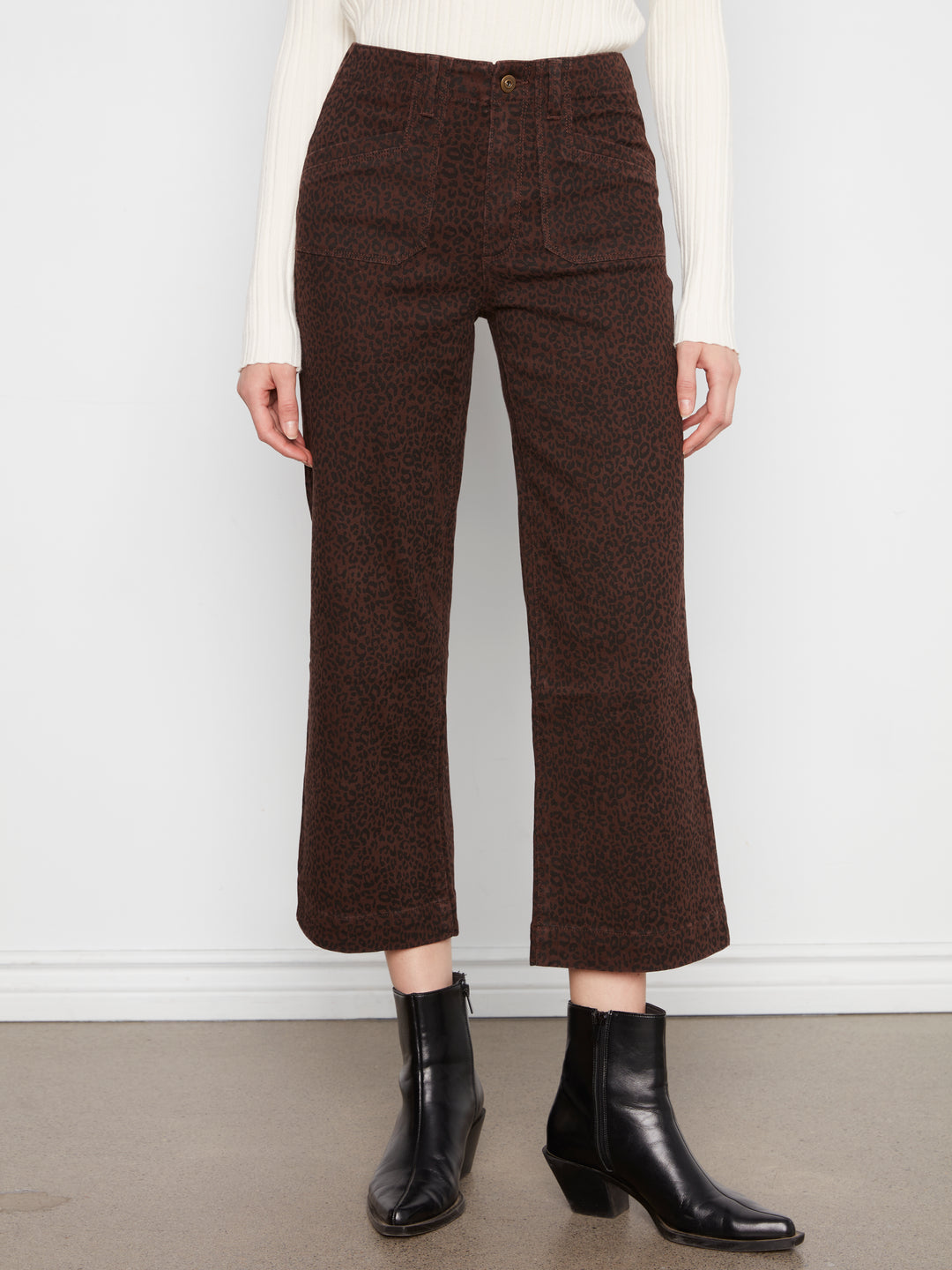 A model is wearing dark brown cropped wide-leg trousers with a textured tweed-like finish, paired with a white ribbed sweater and black ankle boots, standing against a white wall.