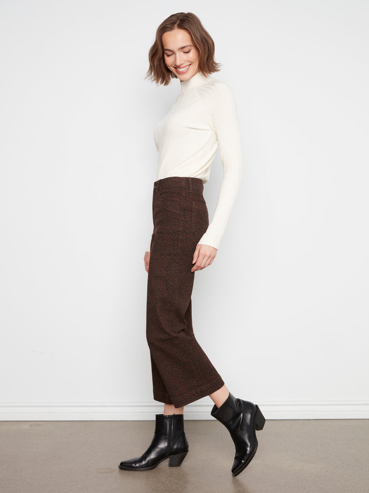 A woman modeling a white long-sleeve mock neck top, a dark brown textured midi skirt, and black ankle boots, standing against a plain white wall.