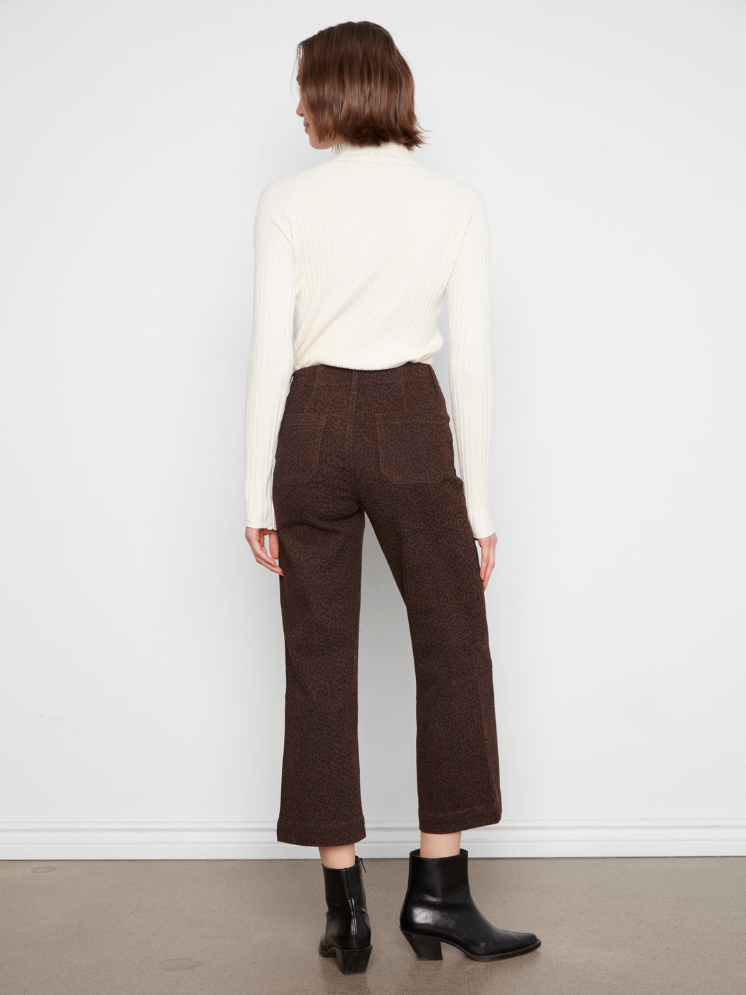 A model shown from the back wearing dark brown cropped trousers, a cream ribbed turtleneck sweater, and black ankle boots against a white studio wall.