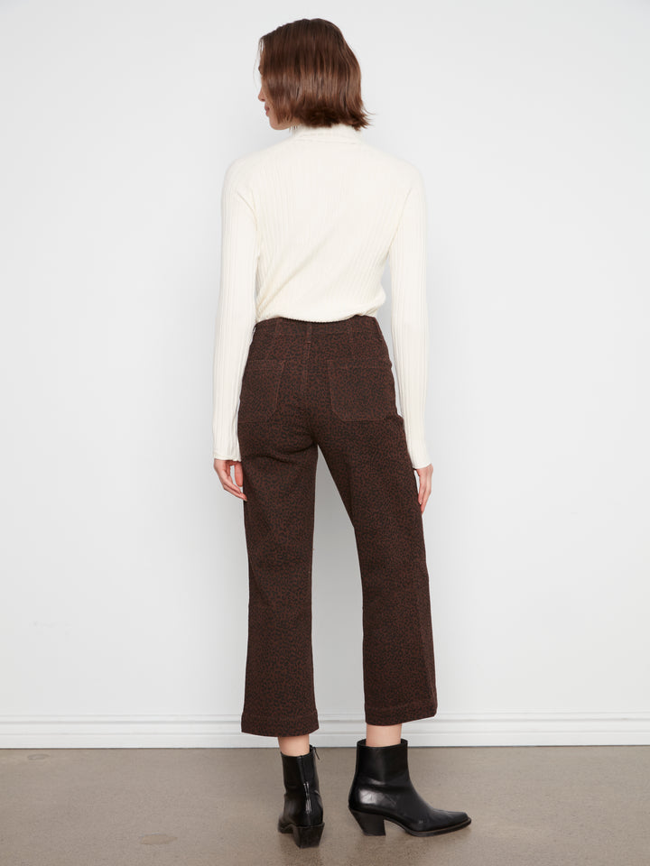A model shown from the back wearing dark brown cropped trousers, a cream ribbed turtleneck sweater, and black ankle boots against a white studio wall.