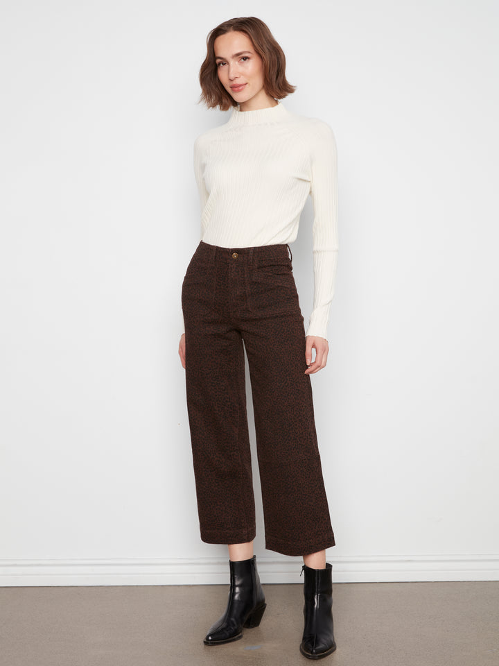 A woman modeling a cream mock-neck long-sleeve top and dark brown cropped wide-leg trousers with black ankle boots, standing against a plain white wall.