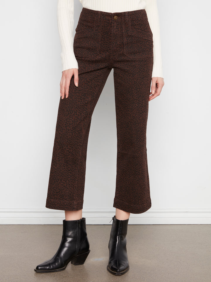 A model is shown from the waist down wearing dark brown cropped wide-leg trousers, a white ribbed sweater, and black leather ankle boots against a white wall and gray floor.