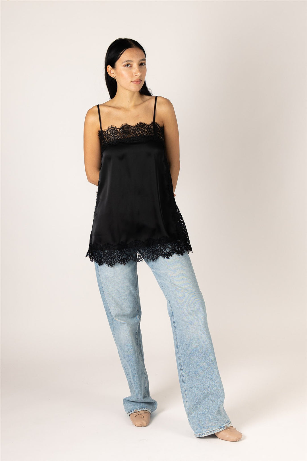 A woman modeling a black satin camisole top with lace trim, paired with light-wash jeans, against a plain light beige studio background.