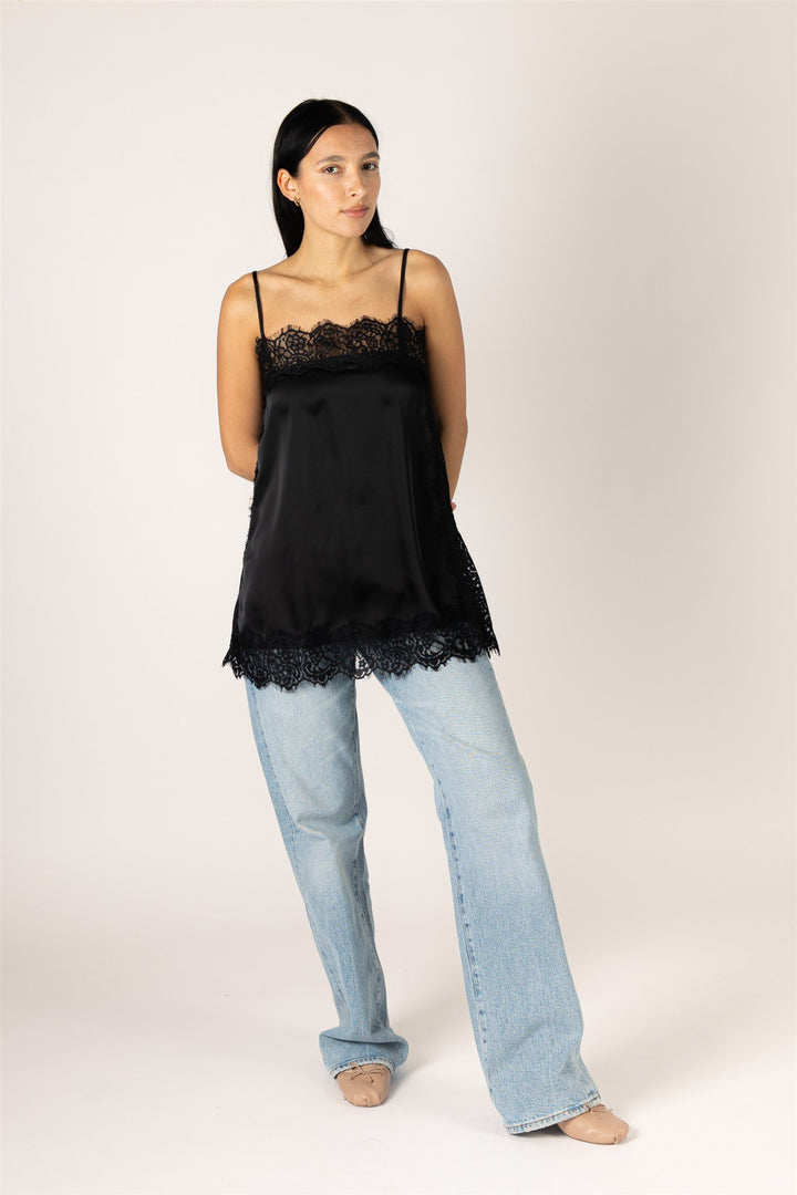 A woman modeling a black satin camisole top with lace trim, paired with light-wash jeans, against a plain light beige studio background.