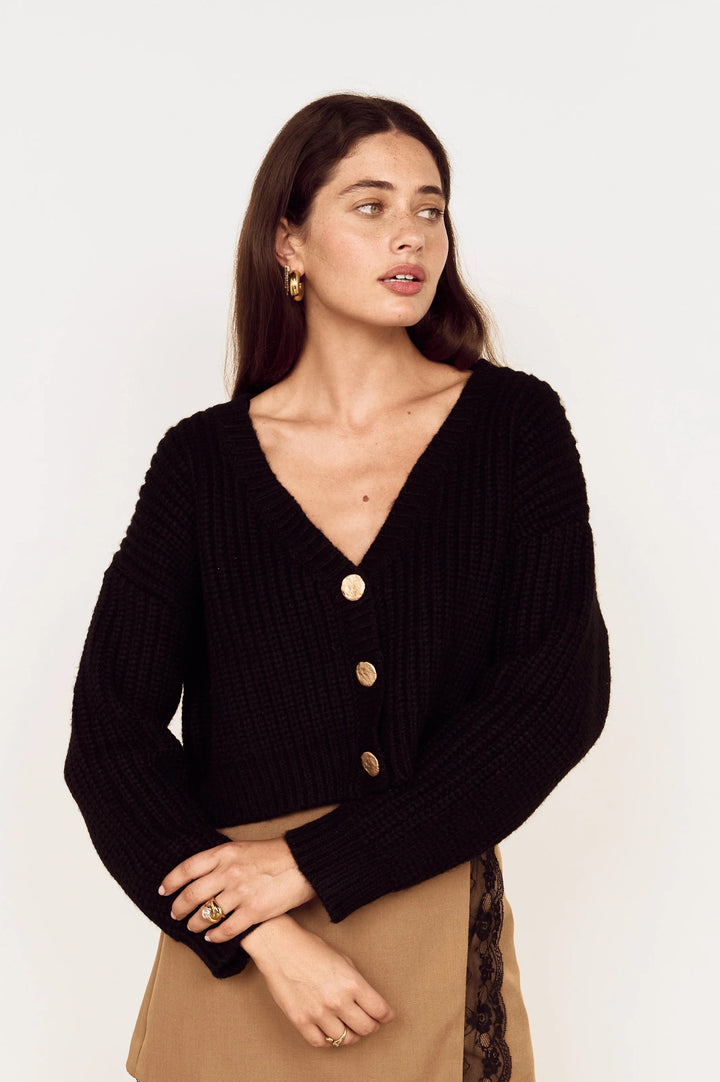 A woman modeling a black ribbed knit cropped cardigan with gold buttons, paired with a tan skirt, against a plain white studio background.